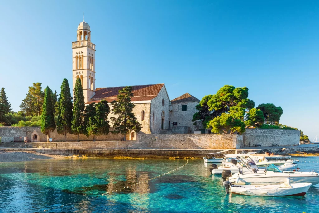 Acqua turchese della baia del mare Adriatico sull'isola di Hvar con monastero francescano e barche nella regione della Dalmazia, Croazia, al sorgere del sole